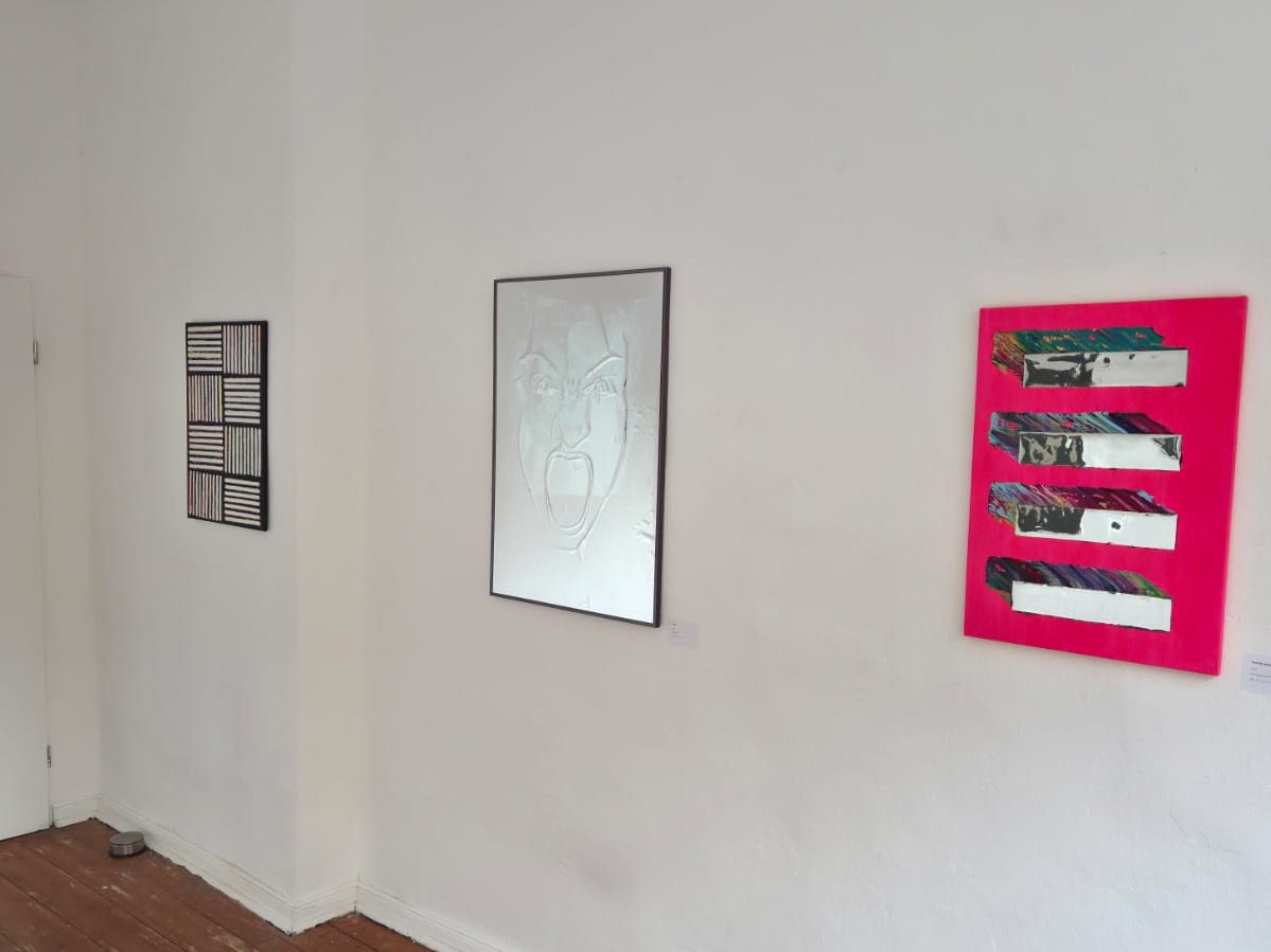 Drei Arbeiten von Joana Diez an der weißen Galeriewand: ein schwarz-weißes Streifenbild, ein weißes Relief mit schreiendem Gesicht, eine pinke Leinwand mit Spiegelstreifen
